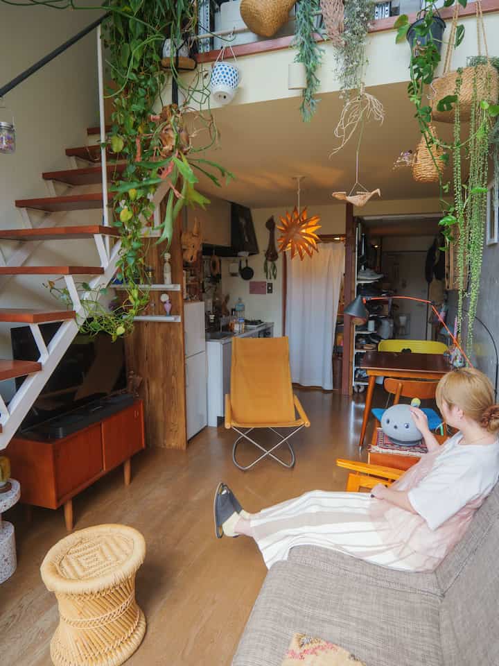 ウッドトーンとナチュラルカラーが調和したワンルームのリビング、天井や壁には多くの植物が吊り下げられ、落ち着いたソファとスツールが配置された雰囲気のあるインテリア