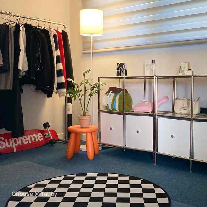 ブルーカーペットの床に黒白のチェッカー柄ラグ、メタルの収納棚と洋服ラック、オレンジのスツールとフロアランプが配置されたモダンな空間