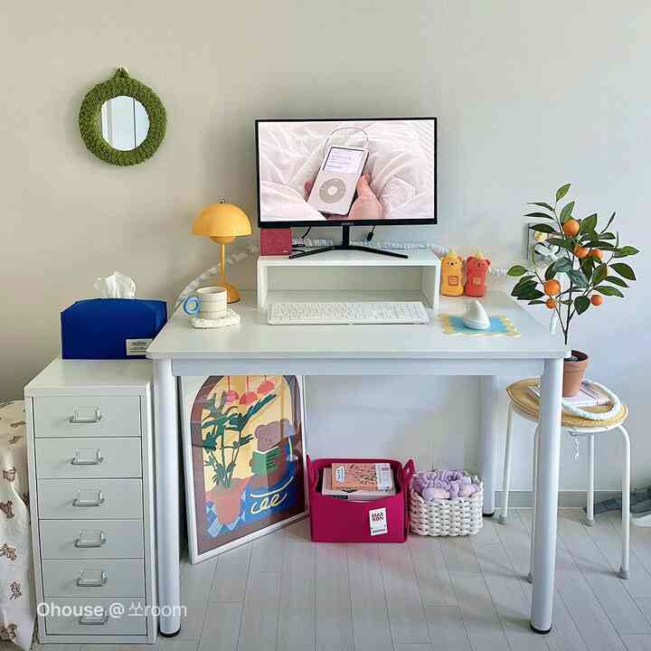 白を基調としたホームオフィス空間で、中央にモニターとデスクが配置され、横に植物と額縁があり、清潔感のある雰囲気のインテリア