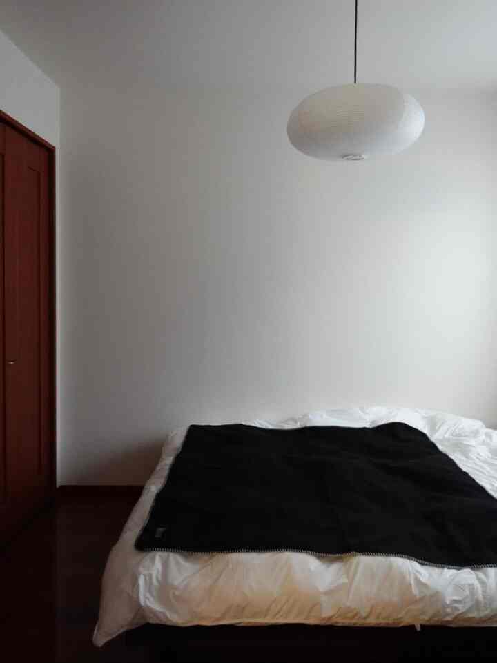 Simple bedroom with white walls and wood tone floor, featuring a centrally placed bed and oval rice paper pendant light