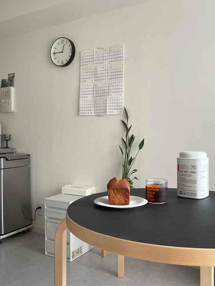 White-walled 1K space featuring wood-toned round dining table, wall clock, calendar, and a home cafe vibe with coffee on table