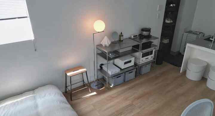Minimal studio apartment with white walls and wood tone flooring, featuring a stool and floor lamp in a simple setting