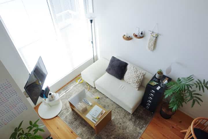 Bright living room with white walls and wood tone flooring featuring a two-seater sofa, vintage rug, coffee table, and plants creating a cozy relaxation space
