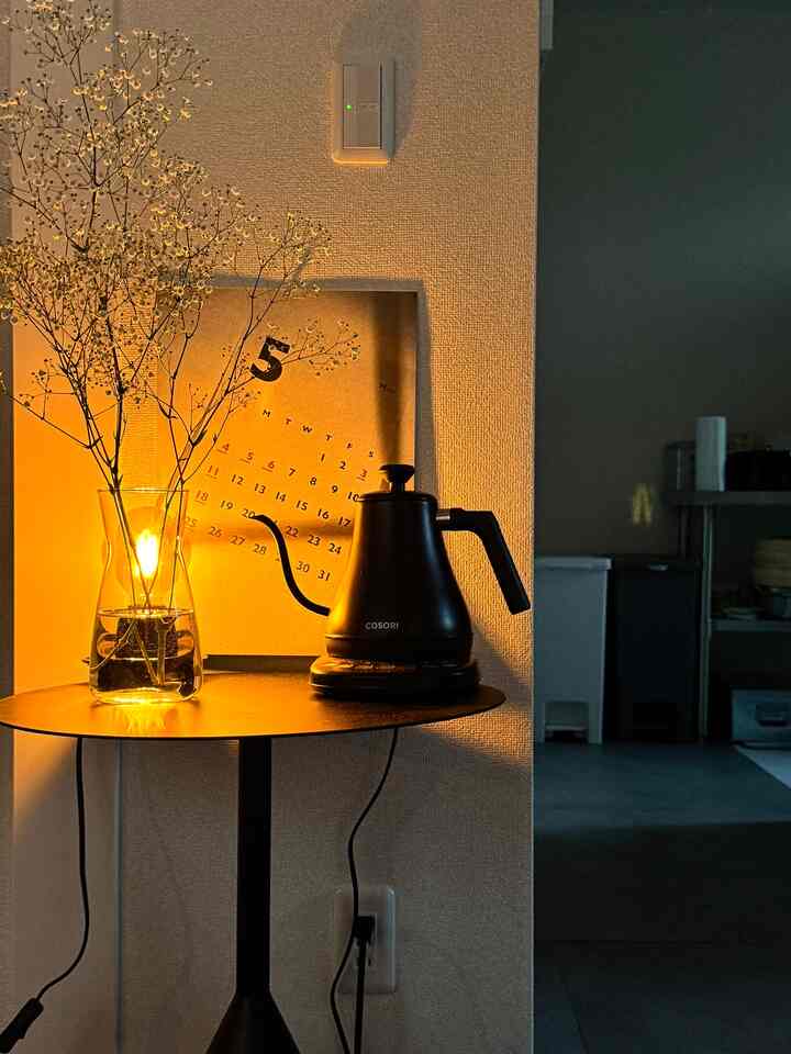 暖かいオレンジ色の光が差し込むキッチンの一角、黒いサイドテーブルに透明な花瓶と電気ポットが置かれた雰囲気のあるインテリア