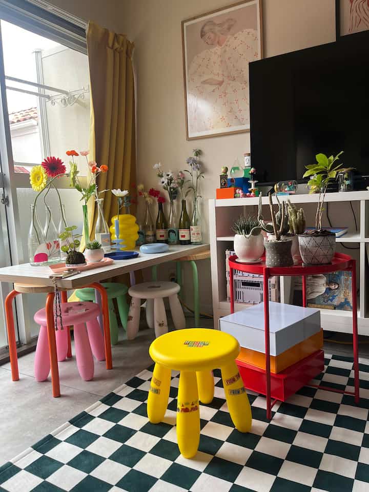 Dining room with yellow curtains and various flower vases featuring colorful stools and checkered rug creating a cozy atmosphere