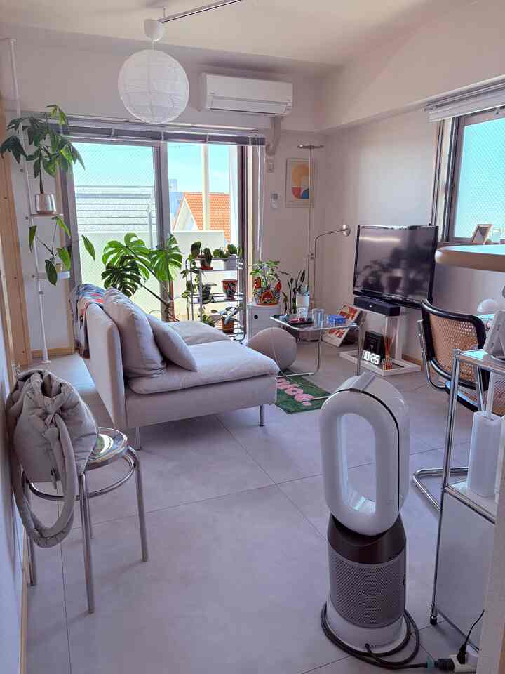 薄いグレーのフロアタイル床とベージュのソファが置かれた1LDKのリビング、複数の植物と自然光が明るく開放的な雰囲気を演出しているインテリア