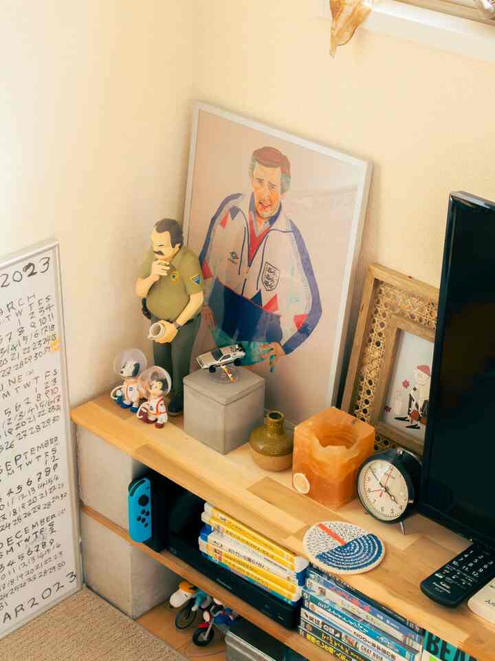 ベージュの壁と木目調の棚がある1LDKのリビングルーム、ミッドセンチュリーモダンのオブジェや時計、ポスターが配置された雰囲気のあるインテリア