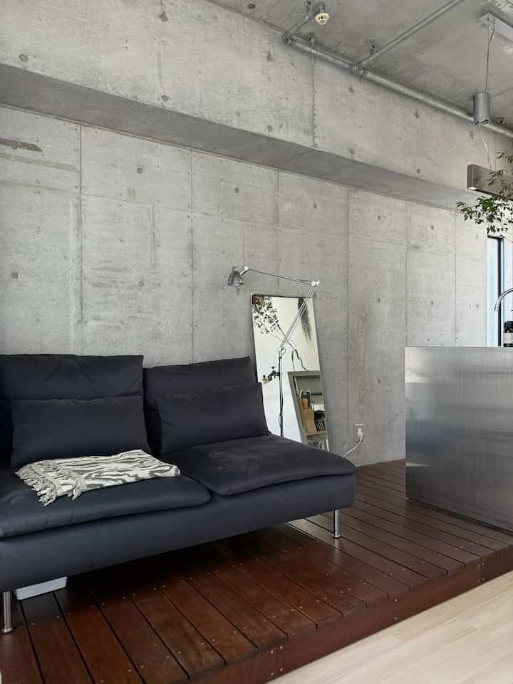 打ちっぱなしコンクリートの壁とステンレスキッチンを備えたモダンなワンルームリビング、ブラックのソファとフロアランプが配置されたシンプルで洗練された空間