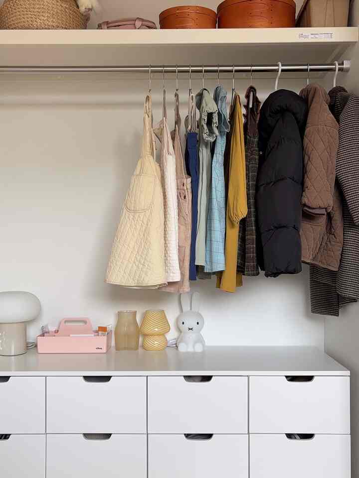 ホワイトトーンの壁とチェストがあるすっきりした収納スペース、様々な服と照明が調和したミニマルなインテリア