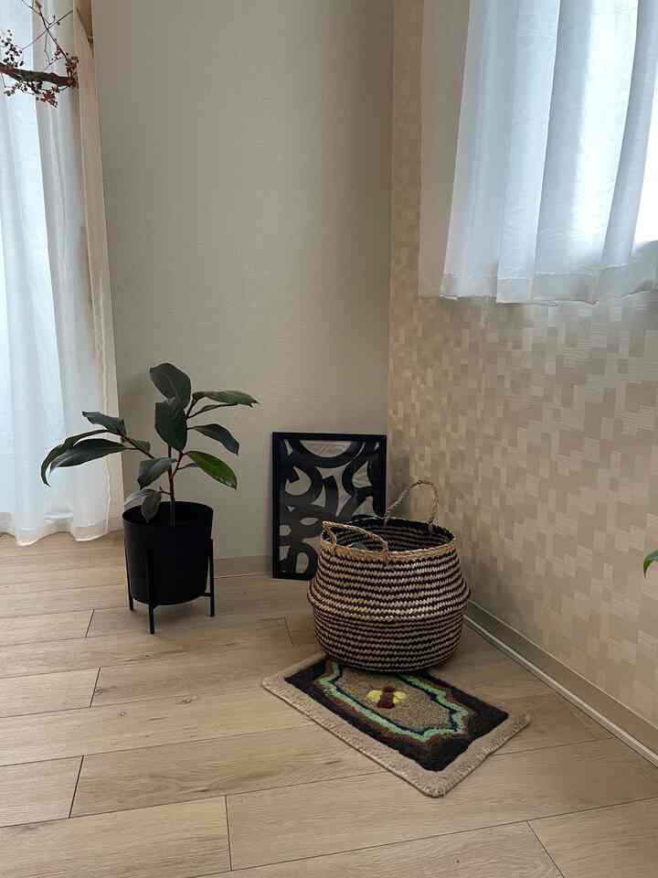 Living room corner with wood tone flooring and beige wallpaper featuring a black plant pot, two-tone woven basket, and a colorful patterned rug creating a cozy atmosphere
