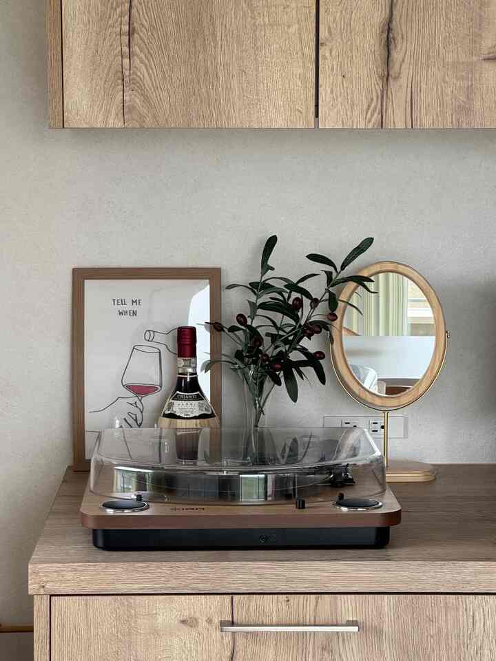 ナチュラルな色調のキッチン、木製キャビネットの上にレコードプレーヤーとワインボトル、ポスターと鏡が配置された家庭的な雰囲気のインテリア