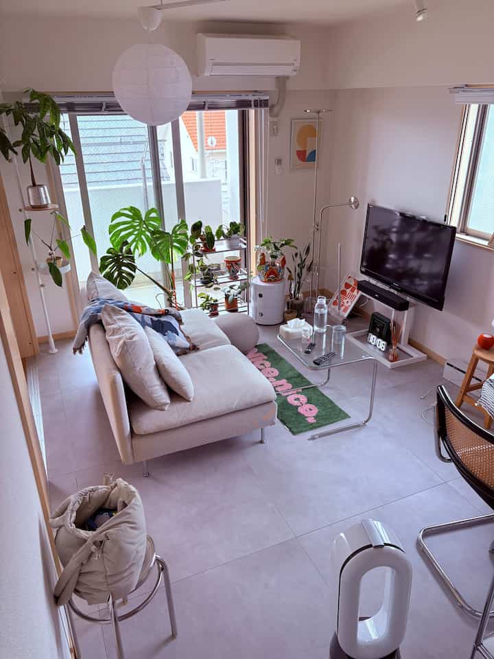 Beige-toned living room featuring a central beige sofa, glass coffee table, and various plants near window creating cozy atmosphere