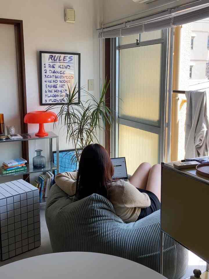 A cozy small studio living room with natural light, featuring a bean bag sofa, orange table lamp, bookshelf, and plants in mid-century modern style