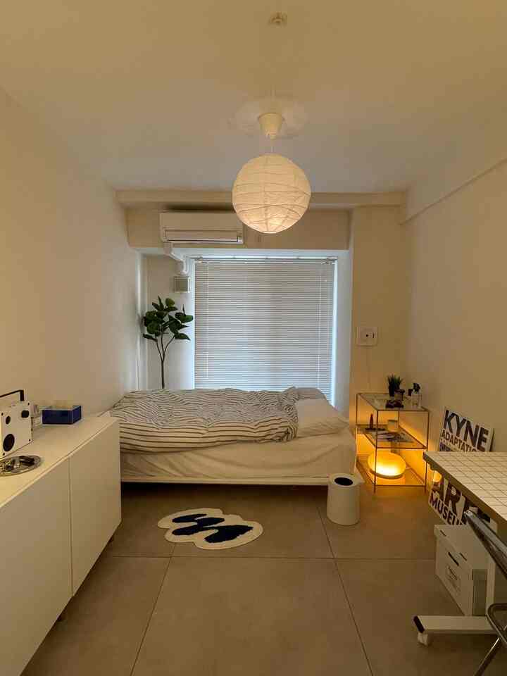 A cozy 1K bedroom with soft beige floor and white walls, featuring modular shelves and warm ambient lighting