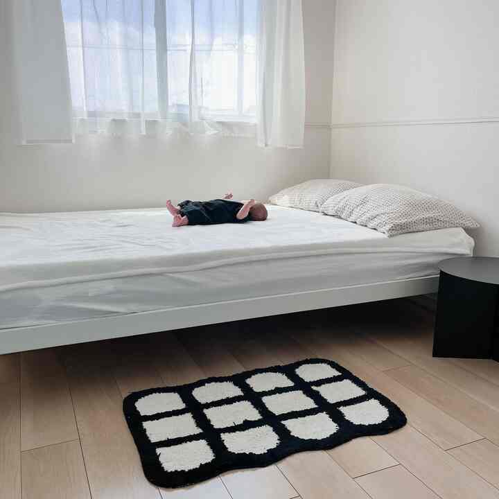 Bright and simple bedroom with white walls and bedding, black side table and rug, featuring a baby lying on the bed