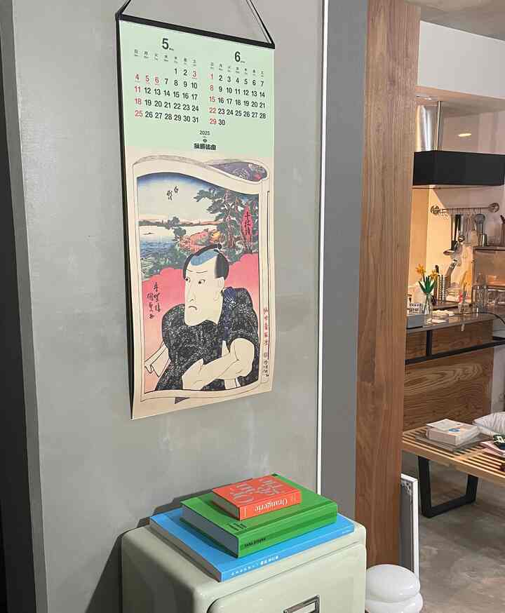 グレーの壁に伝統的な日本画のカレンダーが掛かり、木目調のキッチンとベンチが置かれた内装の1LDK一人暮らし空間