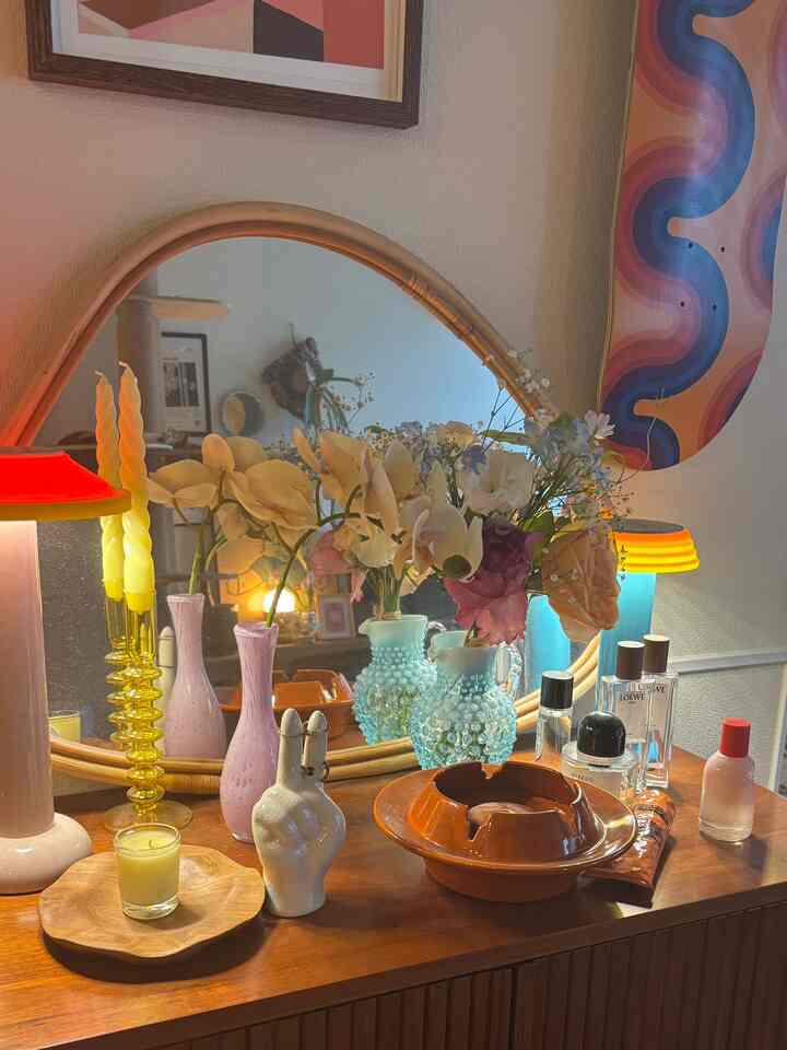 Cozy space featuring brown wood tone cabinet topped with assorted flower vases, candles, objets, and two table lamps near a large mirror