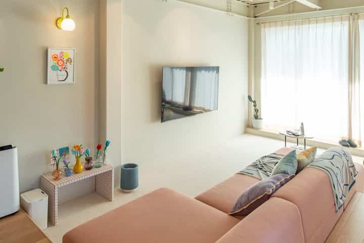 ホワイトとピンクのトーンのリビング、ピンクのソファと壁掛けテレビ、自然光が差し込むミニマルな雰囲気のあるインテリア