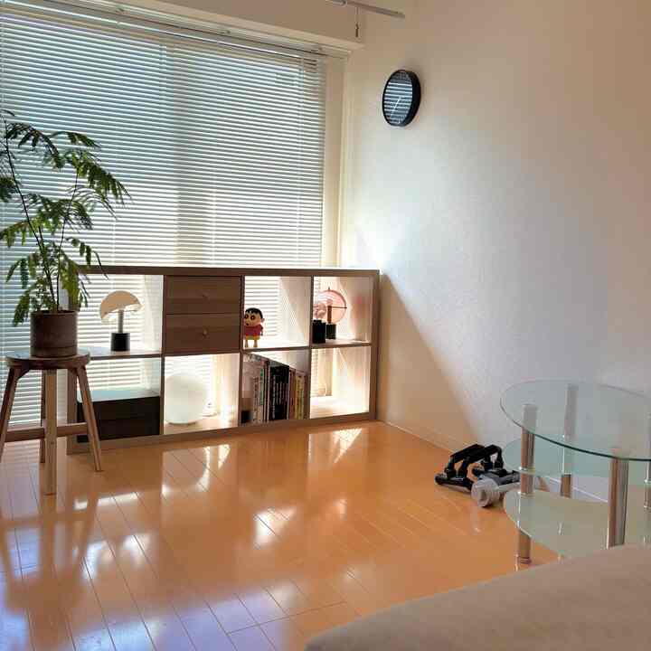 Bright natural-lit living room with white walls and blinds, featuring natural wood shelf, potted plant on wooden stool, and glass table in a clean minimalist space
