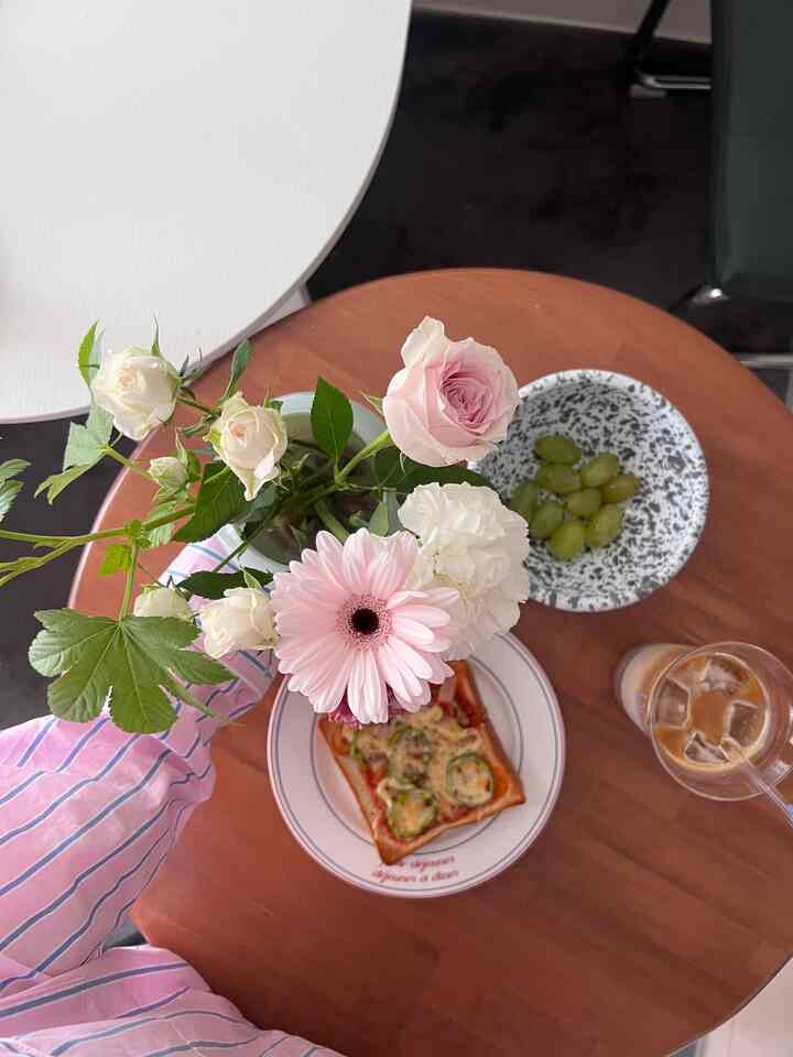 温かみのある木目調のダイニングテーブルにピンクの花瓶とピザトースト、アイスコーヒーが置かれた居心地の良い一人暮らしの空間