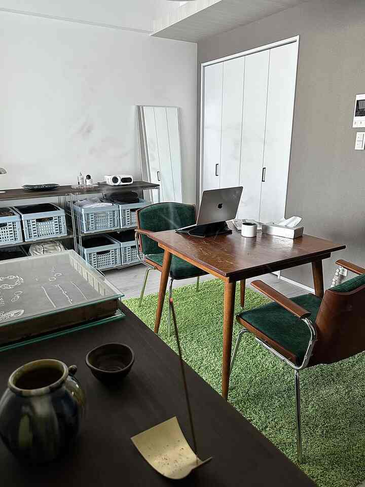 ライトグレーの壁と木目調の家具が配置された1LDKのリビングルーム、緑のラグやチェアと共にモダンなホームオフィスの雰囲気のあるインテリア