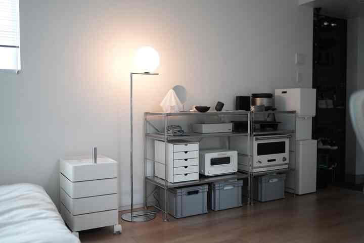 明るい白とメタル素材の棚が配置されたミニマルなキッチンスペース、コンパクトな収納チェストとフロアランプが調和した雰囲気のあるインテリア