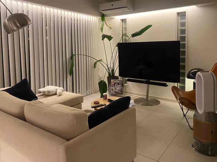 A modern and simple living room in warm beige and white tones featuring a beige sofa, large TV, plants, and vertical blinds