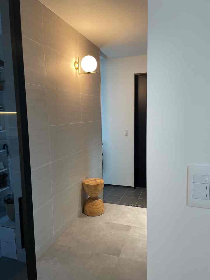 グレーのタイル床と壁面のミニマルモダンな玄関、木目調のスツールと球形の壁照明が配置された清潔感のある空間