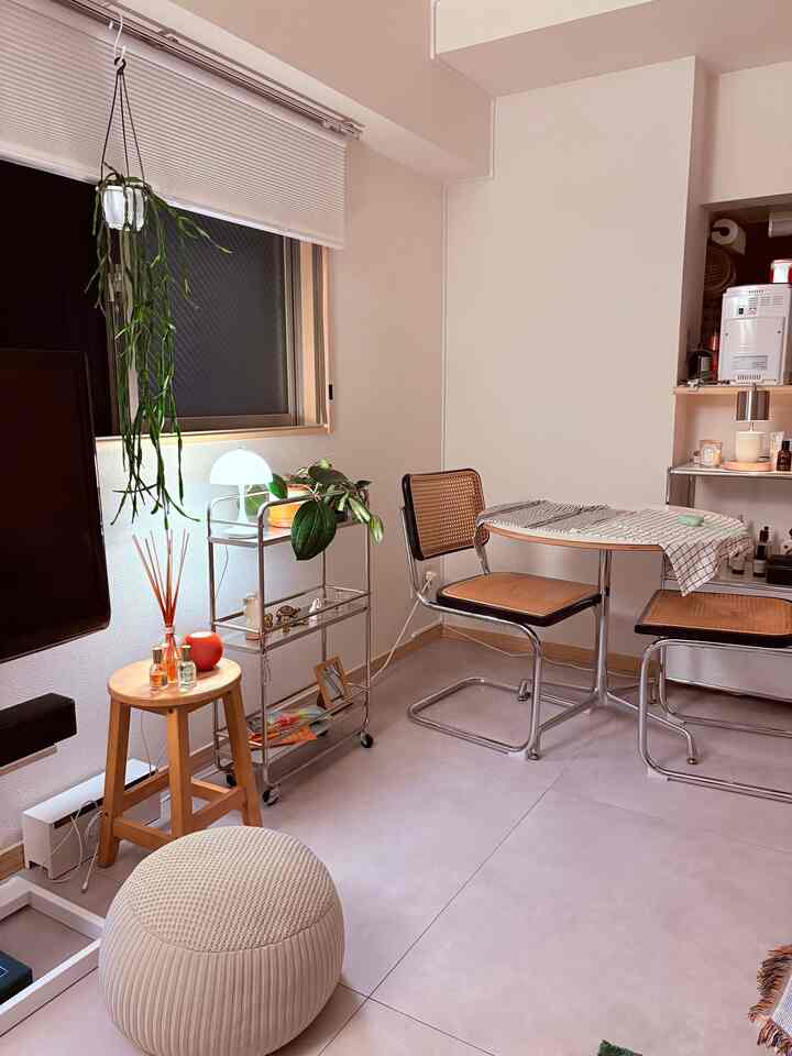 白い壁とライトグレーの床の1LDKリビング、スチールラックとラタンチェア、丸テーブルが配置されたミッドセンチュリーモダンな雰囲気のある空間