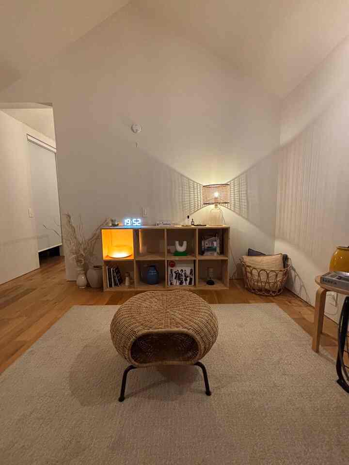 白い壁と木目調のフロアの北欧風リビング、ラタンのスツールとテーブルランプが配置された雰囲気のある空間