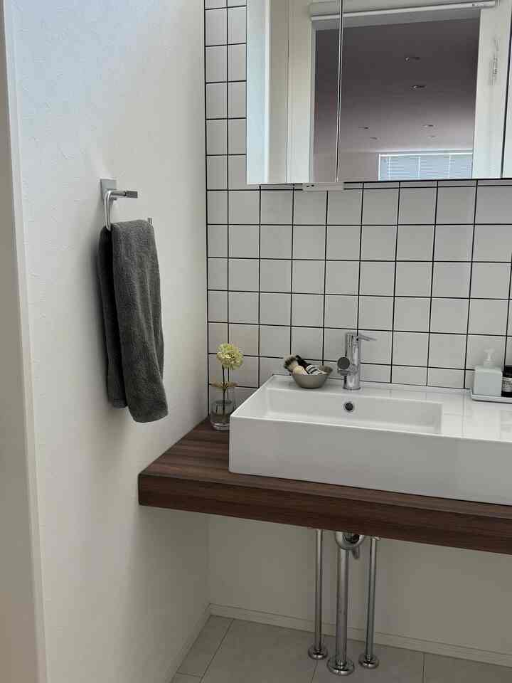 白と木目調のシンプルなバスルーム洗面台、タイル壁と花瓶が配置された清潔感のある空間