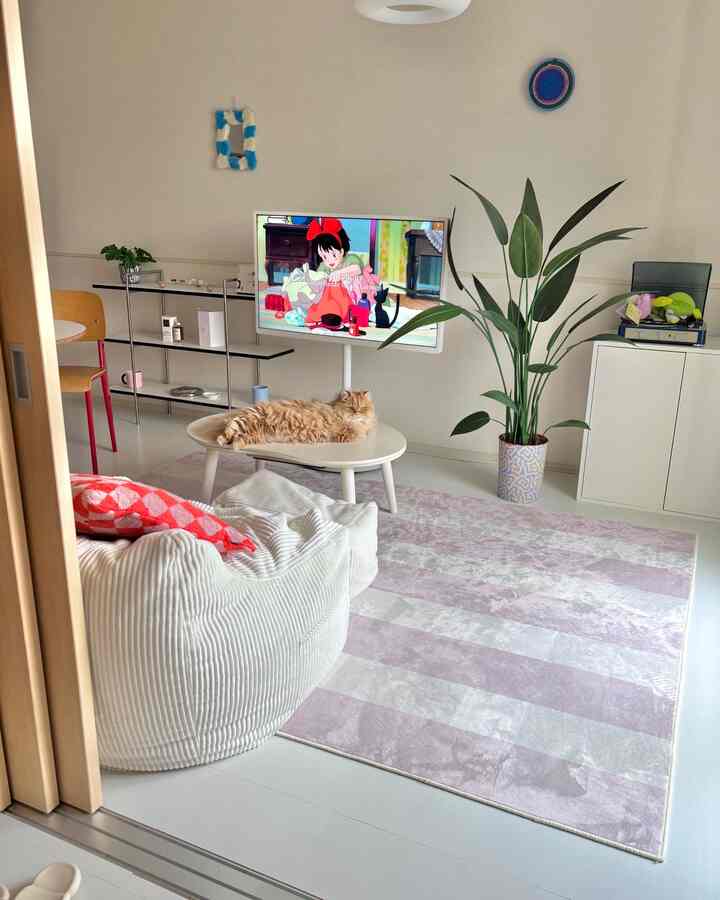 白と薄紫のトーンのリビング、中央の楕円形のテーブルに横たわる猫とビーズクッション、緑の植物が配置された自然な雰囲気のインテリア