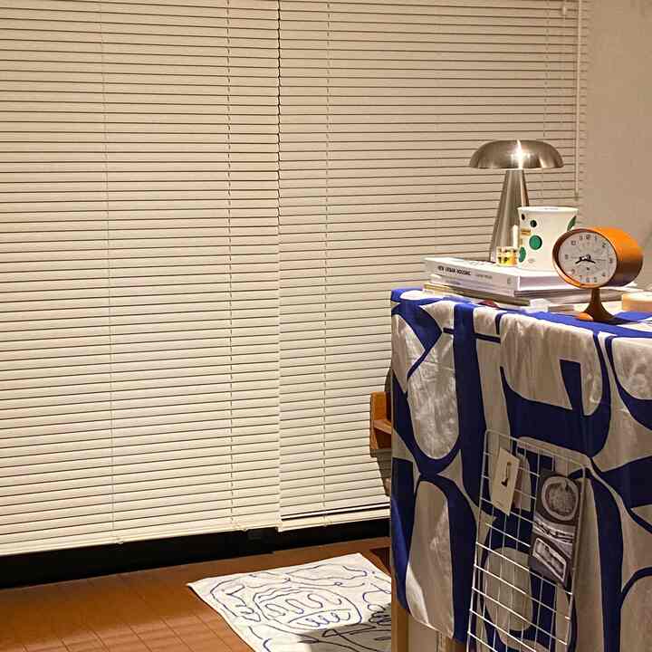 White blinds dominate the background of a single household bedroom featuring a silver mushroom table lamp and wooden clock on a bedside table creating a cozy atmosphere
