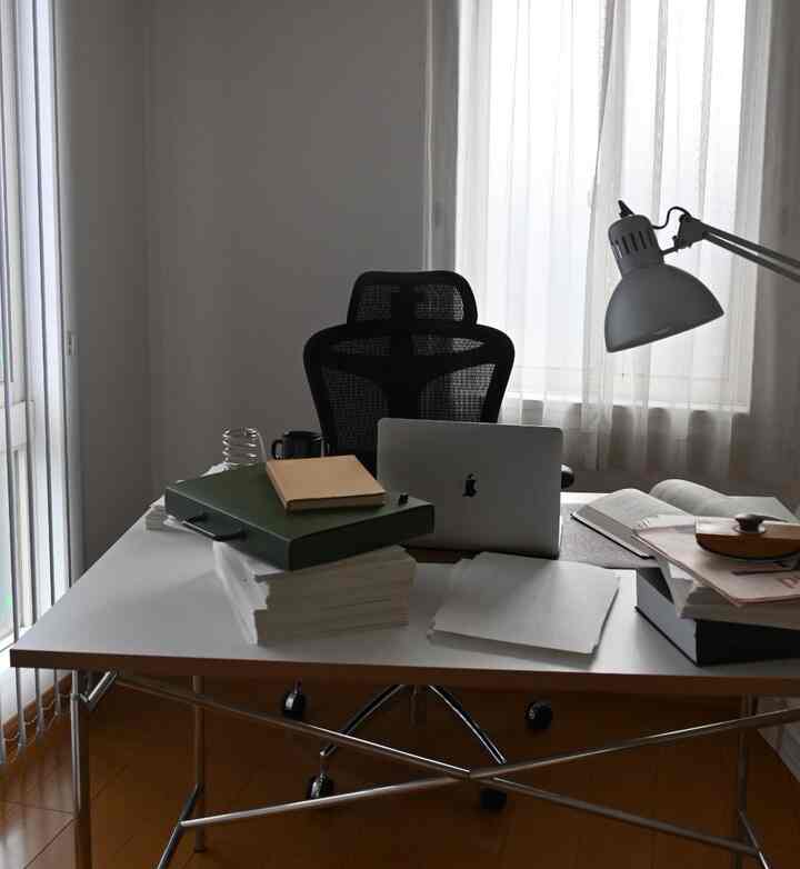 A white and brown toned compact home office featuring a desk with laptop, documents, and a black office chair in a calm study space