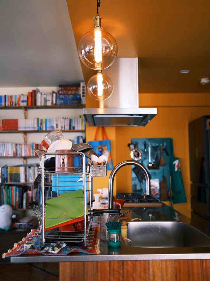 オレンジ色の壁とペンダントライトがある広々としたキッチン、ステンレスシンクと食器乾燥台が配置された雰囲気のある空間