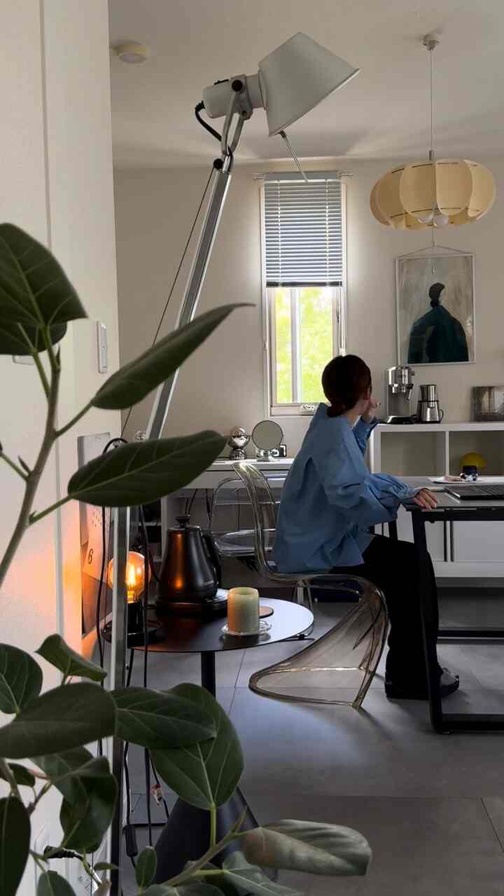 白い壁と茶色の照明があるコンパクトなワンルーム、透明な椅子とデスク、フロアランプが配置された雰囲気のある作業空間