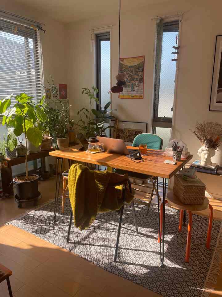 Warm wood tones and natural light fill a 1LDK living room featuring plants and a home office setup for a cozy atmosphere