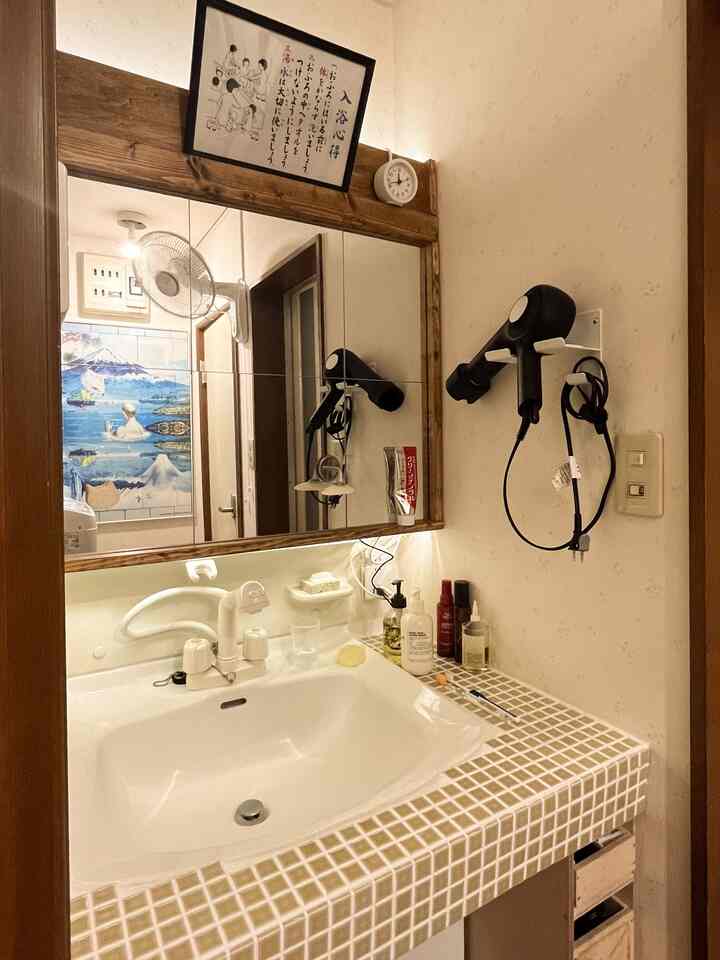 白と木目調が調和した小さなバスルームの洗面スペース、鏡と壁には時計とヘアドライヤーホルダーが配置された実用的なインテリア