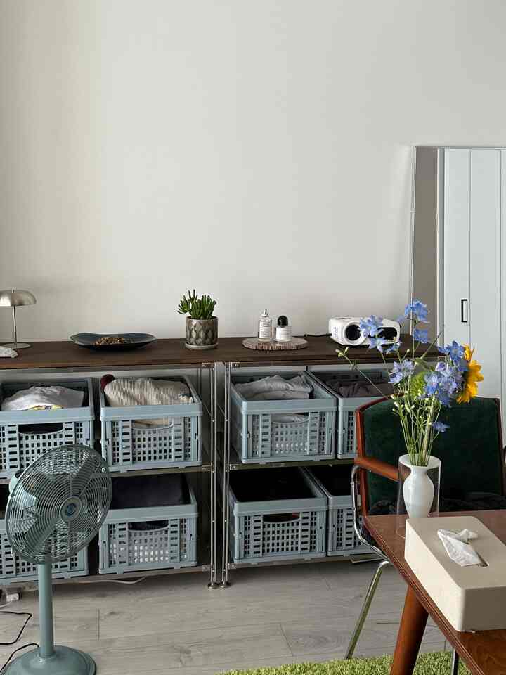 白い壁とライトグレーの棚があるダイニング空間、木目調のテーブルと緑のアームチェア、植物と扇風機が配置されたシンプルなインテリア