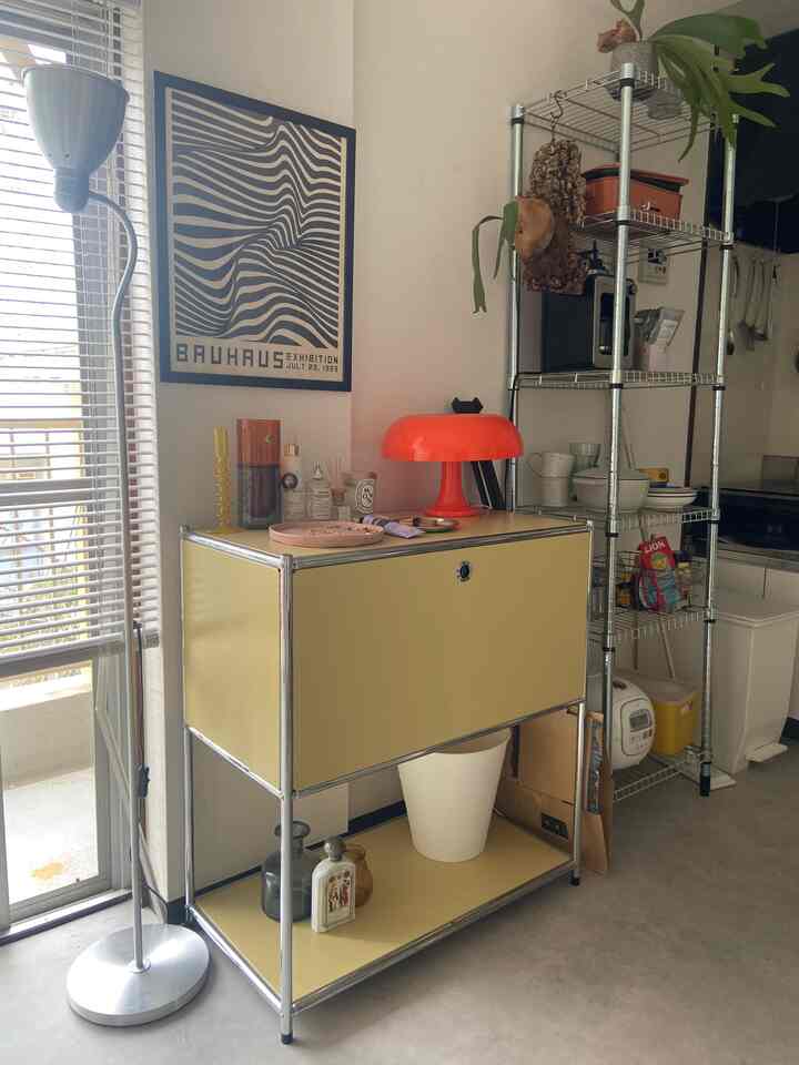 Bright white walls with a mustard yellow side table, kitchen metal shelving, and an orange table lamp create a cozy well-lit space