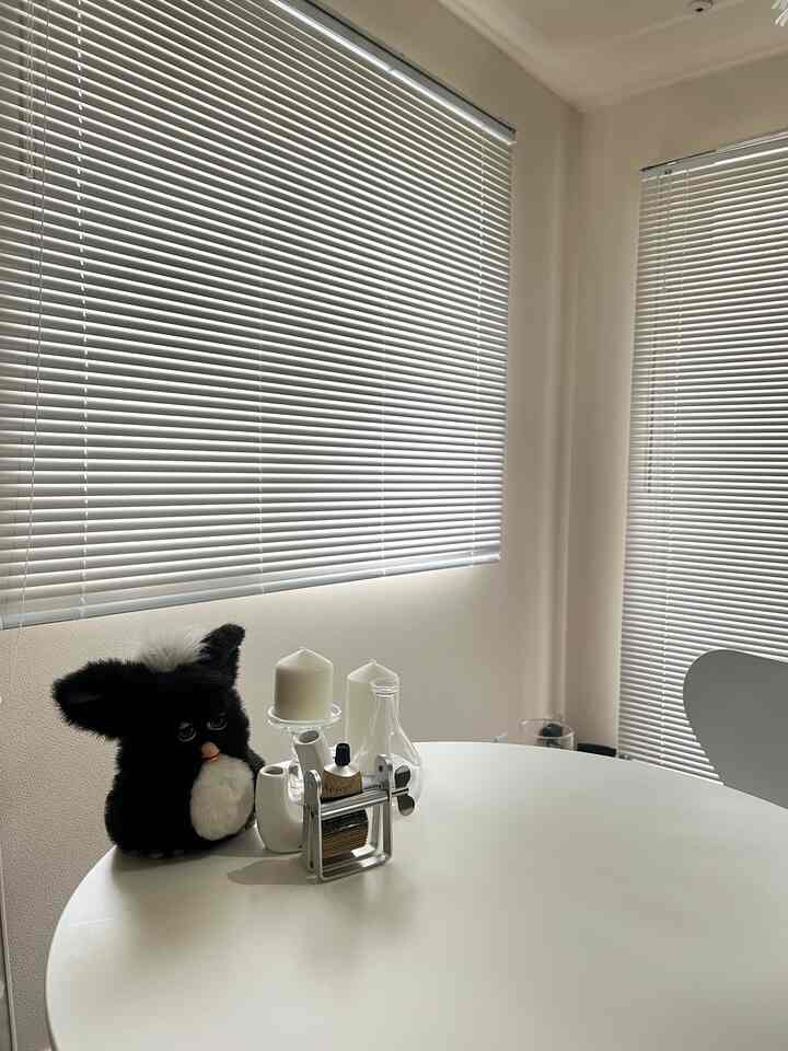 White-toned studio space featuring large windows with white aluminum blinds and a round cafe table with candles, creating a cozy interior