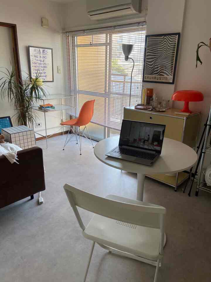 Bright white toned living room featuring a round table with laptop, orange chair, yellow cabinet with floor lamp creating a cozy home office atmosphere