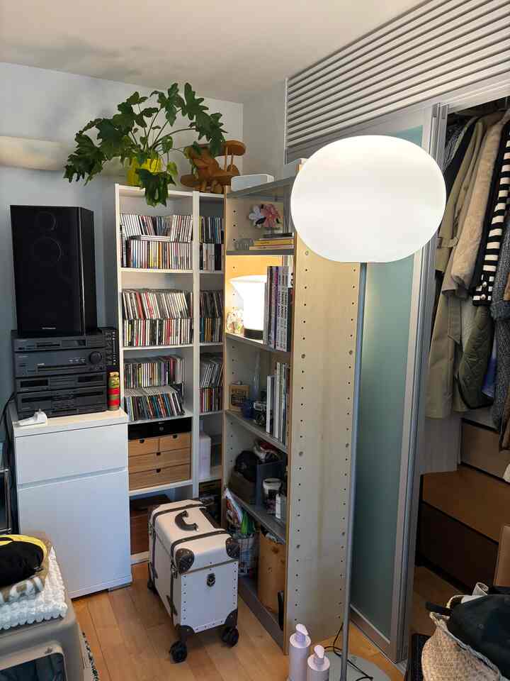 In a compact room with white and wood tones, a floor lamp stands beside bookshelves filled with CDs and a stacked audio system, creating a functional music space