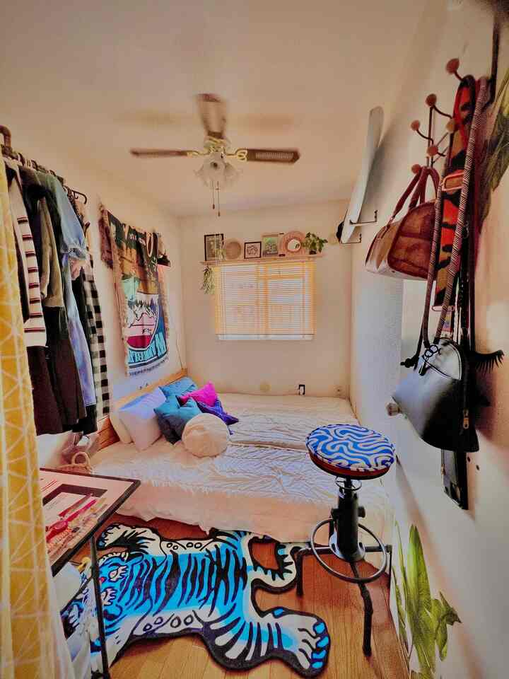 温かみのあるベージュ色調のコンパクトな小部屋、中央にマットレスベッドとブルーのトラ柄ラグ、壁にはバッグが掛けられた印象的な空間