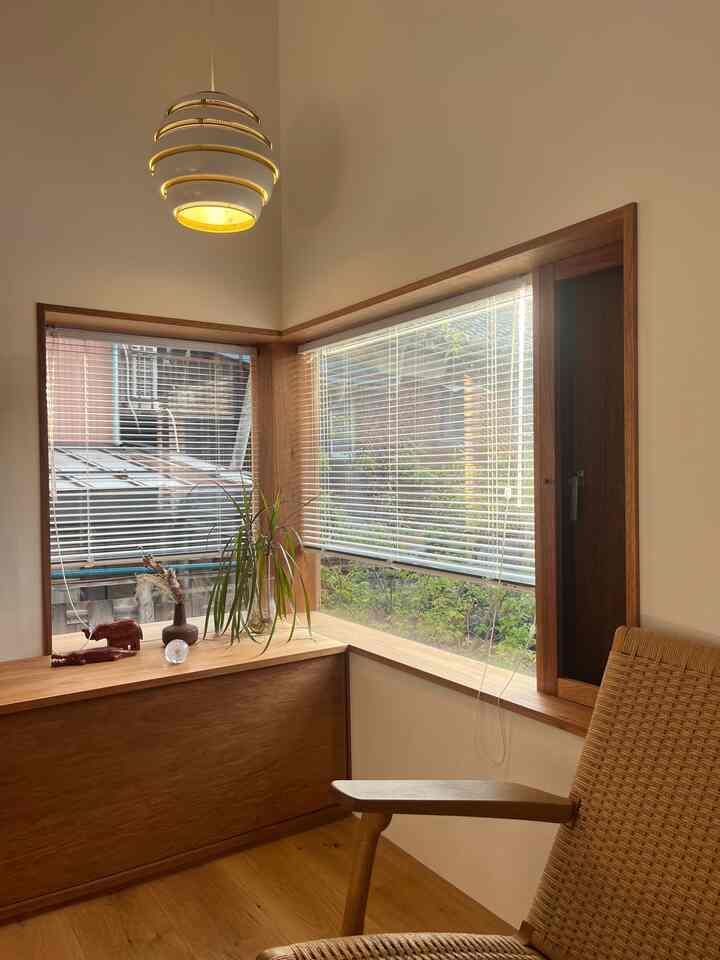 暖かいホワイトと木目調が調和するリビングのコーナー、ブラインドとアームチェア、ペンダントライトが印象的なナチュラルモダンな空間