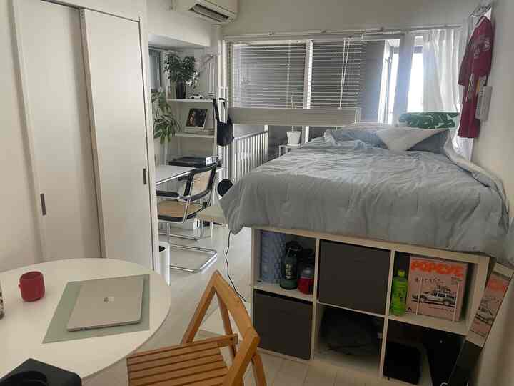 Bright white and light gray toned 1K studio apartment featuring a high bed, desk, and smart storage solutions