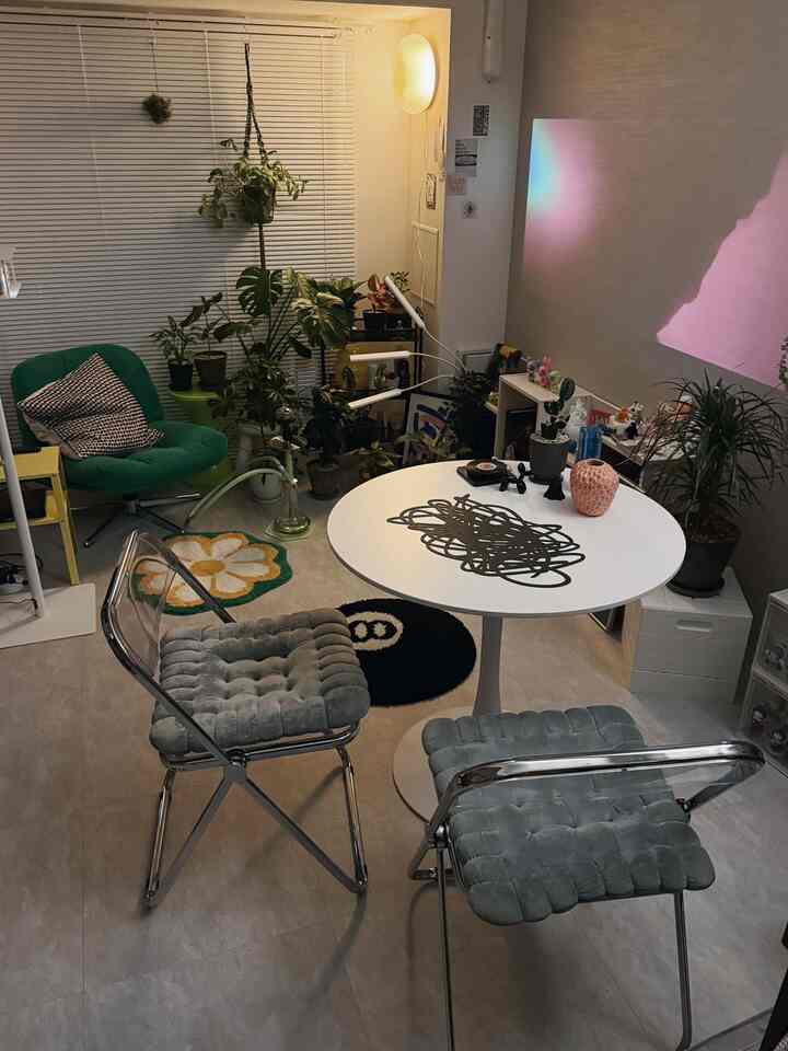 ナチュラルな色調の小さな二人暮らし用リビング、丸い白いテーブルと透明な椅子、緑のアームチェア、多数の植物が配置された温かみのある空間