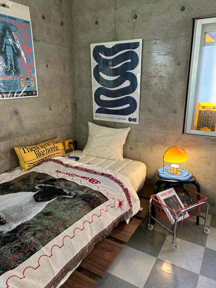 Modern studio bedroom with gray concrete walls and wood-tone bed frame, featuring posters and warm yellow lighting creating a cozy atmosphere
