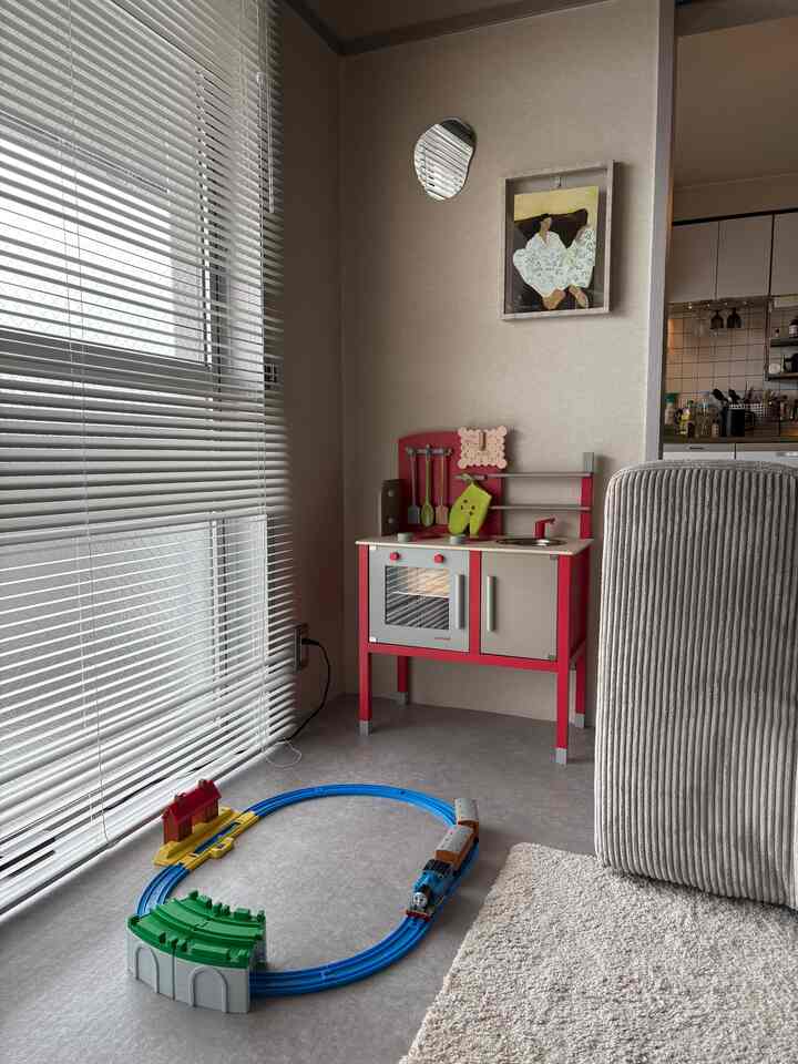 ベージュの壁と大きな窓にブラインドが設置されたコンパクトな子供の遊び場、赤い木目調のキッチンおもちゃとグレーの床に遊び用トラックが配置された印象的な空間