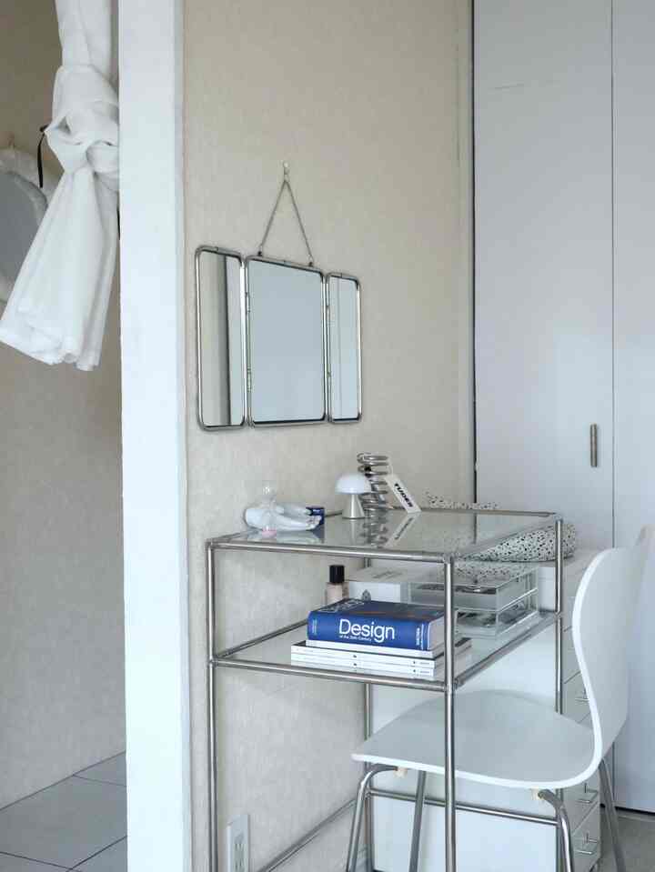 White and metal-toned dressing room corner featuring a glass desk, three-panel mirror, and a blue book accent in a simple space